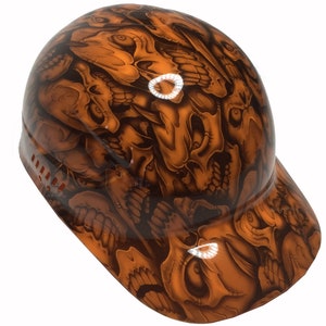 Puede incluir: Un casco de seguridad con un patrón de calavera en tonos marrones y naranjas. El casco tiene una visera y un sistema de ventilación de malla.
