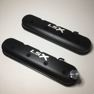 May include: Two black valve covers with the text "LSX" in white lettering. The valve covers have a matte finish and are designed for a car engine.
