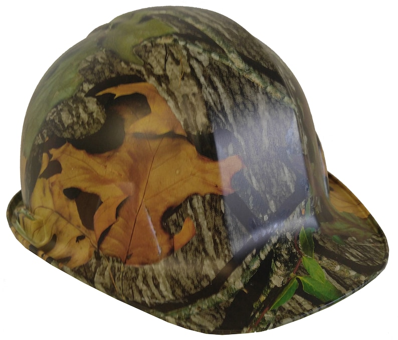 May include: A hard hat with a camouflage pattern featuring brown, green, and tan colours. The pattern includes leaves and tree bark.