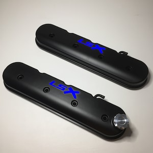 May include: Two black valve covers with blue "LSX" lettering. The valve covers have a smooth, matte finish and are designed for a car engine.