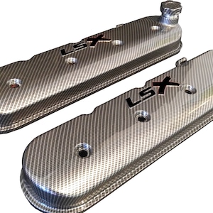 Puede incluir: Un par de tapas de válvulas de motor plateadas con un patrón de fibra de carbono y la inscripción "LSX" en negro.