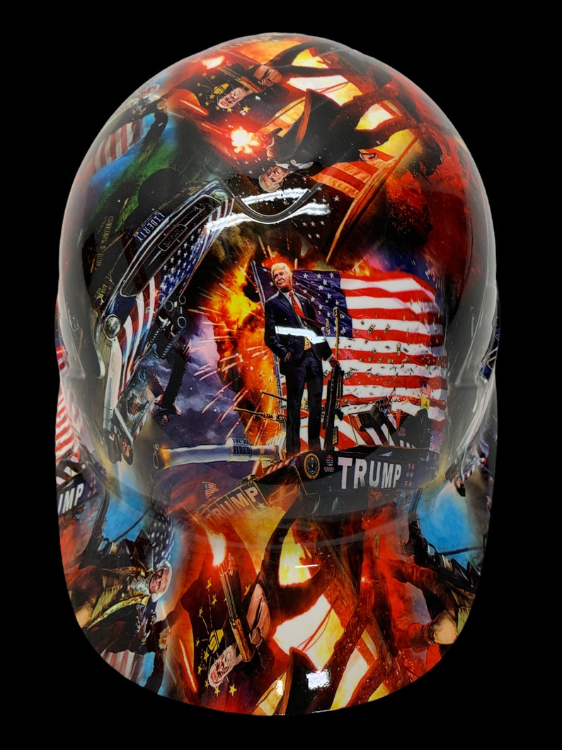 May include: A white hard hat with a colorful graphic design featuring an American flag, a portrait of Donald Trump, and other patriotic imagery. The text "TRUMP" is visible on the hat.