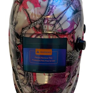 May include: A welding helmet with a camouflage pattern in shades of pink, gray, and black. The helmet has a warning label that reads "Please Remove This Protective Film Prior to Use".