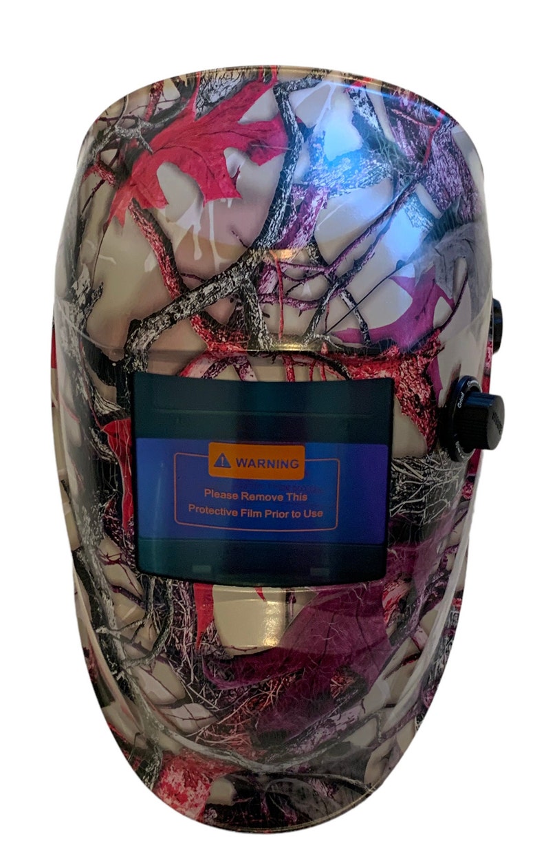 May include: A welding helmet with a camouflage pattern in shades of pink, gray, and black. The helmet has a warning label that reads "Please Remove This Protective Film Prior to Use".