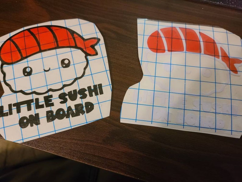 May include: Two vinyl decals featuring a cartoon sushi piece with a red topping and white rice, accompanied by the text "LITTLE SUSHI ON BOARD" in black lettering. The decals are presented on a blue grid transfer paper backing for application.