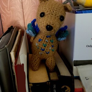 May include: A tan crochet deer plush toy featuring grey antlers, black bead eyes, and blue and teal wings attached to its back. The body of the deer is decorated with a pattern of small blue and teal yarn stitches on its chest and sides.