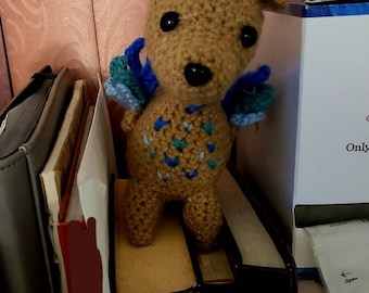 May include: A tan crochet deer plush toy featuring grey antlers, black bead eyes, and blue and teal wings attached to its back. The body of the deer is decorated with a pattern of small blue and teal yarn stitches on its chest and sides.