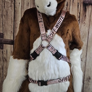 May include: A brown and white furry costume with a horse-like head. The costume features a harness with the text "OFF-WHITE" and "5,400 lbs" printed on the straps. The harness has black buckles and a pink background.