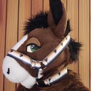 Può includere: Un costume da testa di cavallo marrone con muso bianco e occhi verdi. Il costume presenta un'imbracatura fantasia con impronte di zampe in nero, beige e blu. Lo sfondo è un muro di legno.