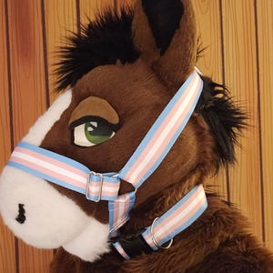 May include: A plush horse head with a brown body, black mane, and white muzzle. The horse head features a halter with blue, pink, and white stripes. The horse has a green eye and a black nose.