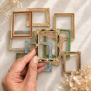 May include: A collection of small, rectangular picture frames featuring ornate, raised gold-tone detailing along the edges. The frames are finished in various colours, including red, blue, green, black, and pink, with a weathered, textured appearance.