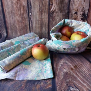 May include: Four reusable cloth produce bags with a green, purple, and yellow marbled pattern. The bags are folded and lying on a wooden surface. A fifth bag is filled with red apples and is sitting on the wooden surface.