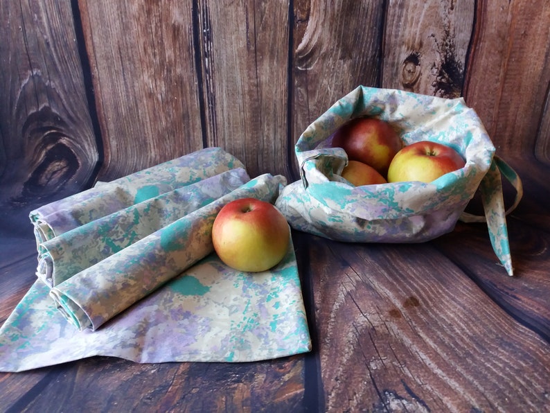 May include: Four reusable cloth produce bags with a green, purple, and yellow marbled pattern. The bags are folded and lying on a wooden surface. A fifth bag is filled with red apples and is sitting on the wooden surface.