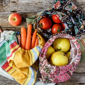 May include: Four reusable produce bags filled with fresh produce. A red apple, carrots, tomatoes, and yellow apples are visible in the bags. The bags are made of fabric with different patterns.