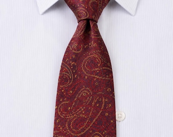 May include: A deep burgundy necktie featuring an intricate, repeating gold and dark red paisley pattern. The tie is knotted and positioned against a crisp, white, vertically textured dress shirt with a visible white button on the right side.