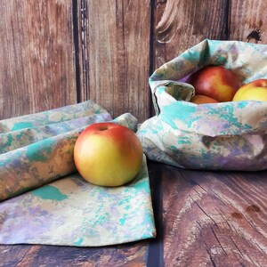 May include: Set of four reusable fabric produce bags with a green, purple, and yellow marbled pattern. The bags are folded and stacked on a wooden surface. A single bag is filled with red apples and is sitting on the wooden surface.