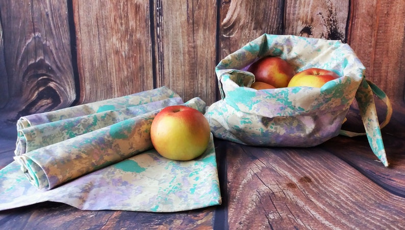 May include: Set of four reusable fabric produce bags with a green, purple, and yellow marbled pattern. The bags are folded and stacked on a wooden surface. A single bag is filled with red apples and is sitting on the wooden surface.