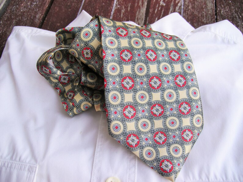 May include: A beige silk tie with a repeating pattern of red and green circles and flowers. The tie is folded and placed on a white shirt.