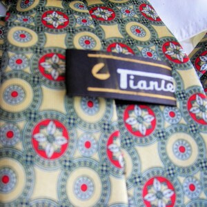 May include: A yellow and green patterned tie with a red and white floral design. The tie has a black label with the word "Tiano" in white lettering.