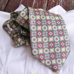 May include: A beige silk tie with a repeating pattern of red and green circles and flowers. The tie is folded and placed on a white shirt.