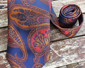May include: A paisley patterned tie with a blue background, brown and orange paisley designs, and red accents. The tie is tied and laid on a wooden surface.