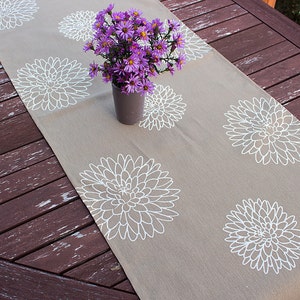May include: A beige table runner with a white floral pattern. The runner is decorated with a bouquet of purple flowers in a gray vase.