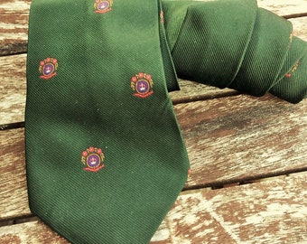May include: A dark green silk tie with a repeating pattern of a crest featuring a crown, flowers, and a ship's wheel.