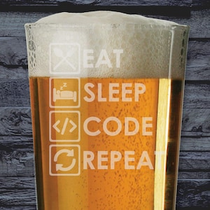 May include: A pint glass filled with beer and foam, with the words "EAT SLEEP CODE REPEAT" etched on the side. The glass sits on a wooden surface, against a dark grey brick wall. The White Elephant logo is visible in the top left corner.