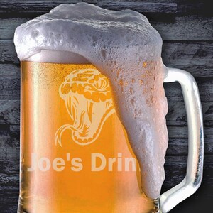 May include: A clear glass beer stein with a snake design and the text "Joe's Drink" etched on the side. The stein is filled with a light-coloured beer and has a thick layer of foam on top.