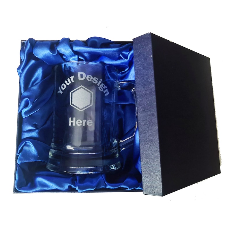 May include: A clear glass beer mug with a handle, in a blue satin lined box. The mug is engraved with the text "Your Design Here" and a hexagon shape.