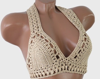May include: A beige crochet halter top featuring a deep V-neckline, intricate openwork stitch patterns, and a textured border along the bottom edge. The garment is constructed with a continuous yarn design that forms a supportive neck strap.