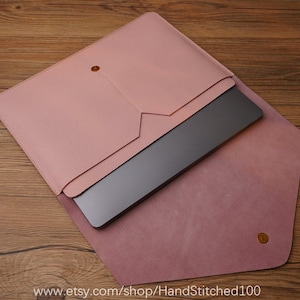 May include: A blush pink leather laptop sleeve with a flap closure and a gold button. The sleeve is open and a laptop is inside.