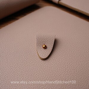 May include: A light pink leather notebook with a gold metal closure. The notebook has a textured surface and the text "www.etsy.com/shop/HandStitched100" is printed on the bottom.