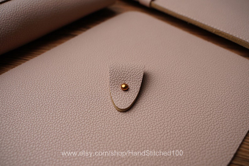May include: A light pink leather notebook with a gold metal closure. The notebook has a textured surface and the text "www.etsy.com/shop/HandStitched100" is printed on the bottom.