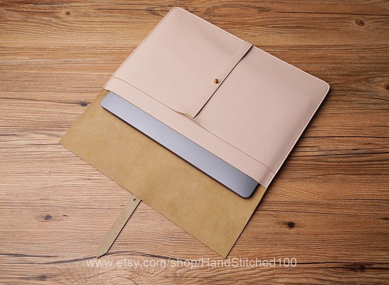 May include: A light pink leather laptop sleeve with a brown suede lining. The sleeve has a flap with a magnetic closure and a small pocket on the front.