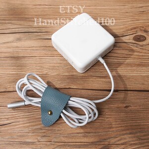 Op de afbeelding: Een witte laptoplader met een grijze kabel is om een ​​blauwe leren snoerorganizer met een gouden knoop gewikkeld. De lader en de snoerorganizer bevinden zich op een houten oppervlak.