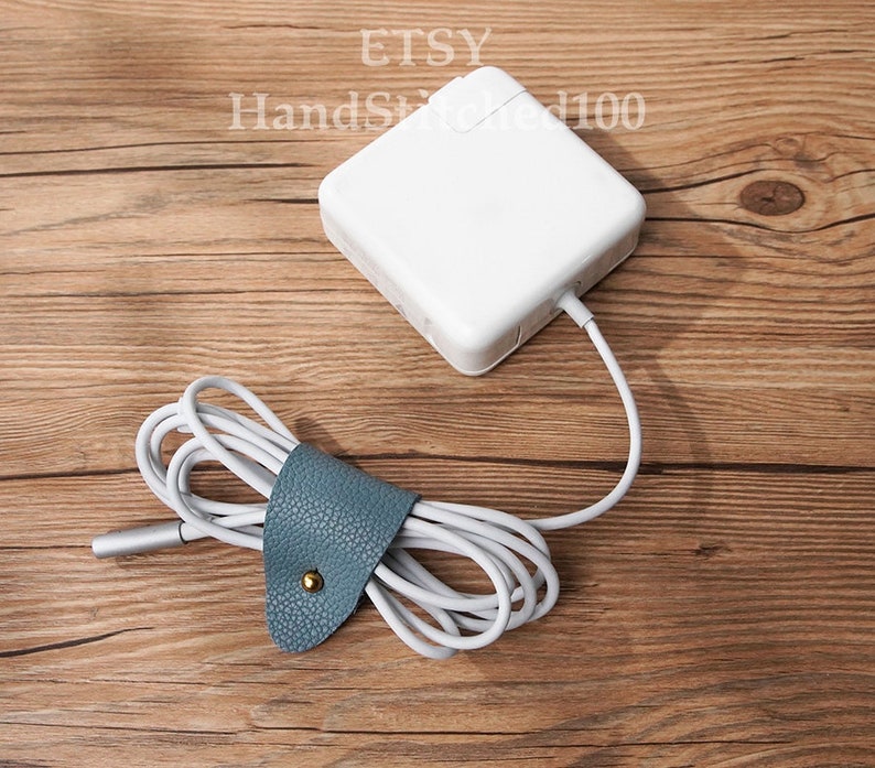 Op de afbeelding: Een witte laptoplader met een grijze kabel is om een ​​blauwe leren snoerorganizer met een gouden knoop gewikkeld. De lader en de snoerorganizer bevinden zich op een houten oppervlak.
