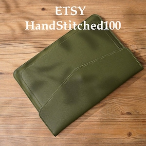 May include: A green leather laptop sleeve with a flap closure and hand-stitched details. The sleeve is on a wooden surface. ETSY HandStitched100