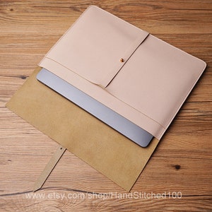 Könnte beinhalten: Eine hellrosa Laptop-Hülle aus Leder mit Knopfverschluss. Ein silberner Laptop ist in der geöffneten Hülle zu sehen. Die Hülle liegt auf einer Holzoberfläche. Der Etsy-Shop-Name, HandStitched100, ist unten links sichtbar.