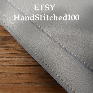Puede incluir: Primer plano de un art&iacute;culo de cuero gris claro con costuras visibles. El texto "ETSY HandStitched100" est&aacute; impreso en blanco. El cuero tiene una superficie texturizada.