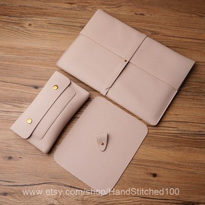 以下が含まれることがあります： 金色のアクセントが付いた薄いピンクの革製のラップトップスリーブ3点セット。一番大きなスリーブはストラップ留め、一番小さなスリーブはポケット付き。スリーブは木製の表面に置かれています。