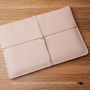 May include: A light pink leather laptop sleeve with a flap closure and a gold button. The sleeve is on a wooden surface.