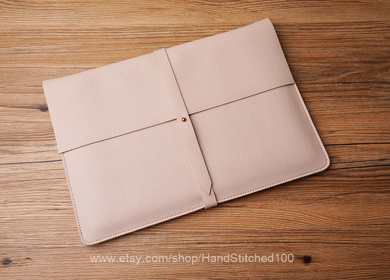 May include: A light pink leather laptop sleeve with a flap closure and a gold button. The sleeve is on a wooden surface.