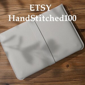 Puede incluir: Una funda para port&aacute;til de cuero gris claro con una costura vertical en el centro. El nombre de la tienda Etsy "HandStitched100" est&aacute; impreso en blanco en la parte superior. La funda est&aacute; sobre una superficie de madera.