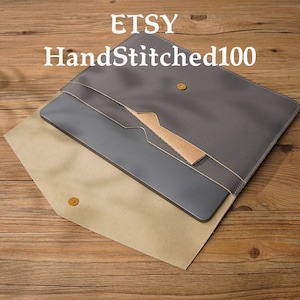 May include: A gray leather laptop sleeve with a beige leather flap and a brown leather pocket. The sleeve has a gold button closure. The text "HandStitched100" is printed on the flap.