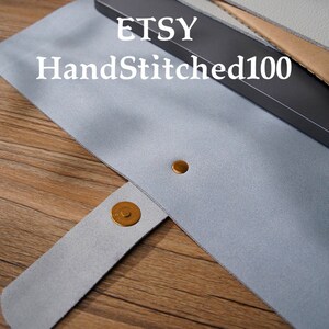 Puede incluir: Un estuche para bol&iacute;grafos de fieltro gris claro con cierre magn&eacute;tico a presi&oacute;n. El estuche tiene un broche dorado y un remache dorado a juego. El texto "ETSY HandStitched100" est&aacute; impreso en el estuche.