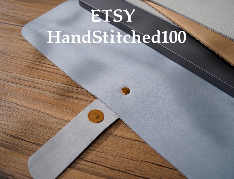 Puede incluir: Un estuche para bol&iacute;grafos de fieltro gris claro con cierre magn&eacute;tico a presi&oacute;n. El estuche tiene un broche dorado y un remache dorado a juego. El texto "ETSY HandStitched100" est&aacute; impreso en el estuche.