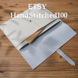 May include: A white leather laptop sleeve with a light blue flap and a gold button closure. The flap has a small hole for a strap. The text "Etsy HandStitched100" is printed on the front of the sleeve.