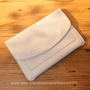 May include: A light beige leather laptop sleeve with a flap closure and a pocket on the front. The sleeve is lying on a wooden surface.