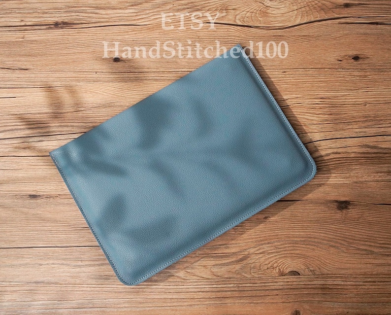 Op de afbeelding: Een lichtblauwe leren laptophuls. De hoes ligt plat op een houten oppervlak.